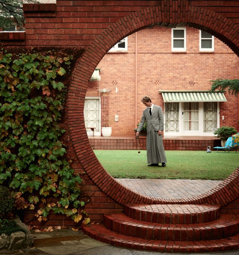 Man in a vintage gray suit standing on a green lawn, holding a croquet mallet, viewed through a circular brick arch with a brick house in the background.