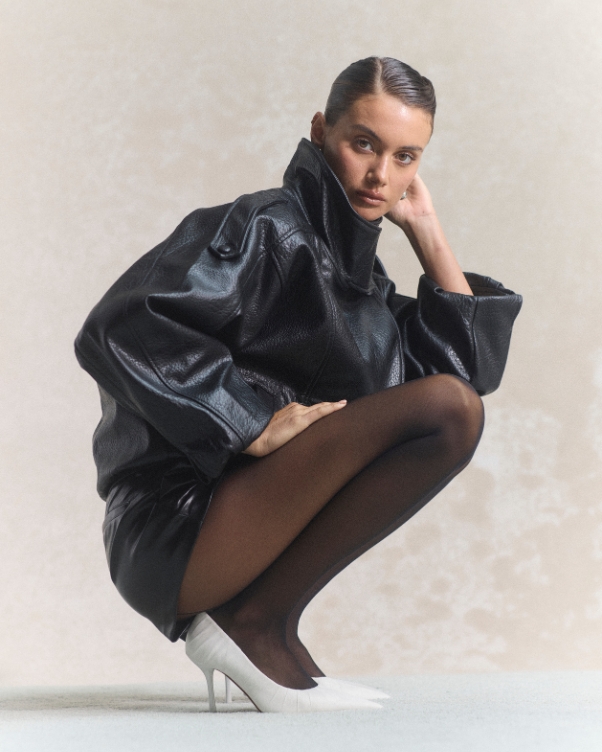 Model crouching in a black oversized leather coat, sheer black tights, and white high heels against a light textured background.