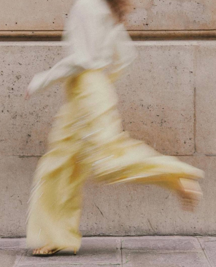Blurry side view of a person in a white top and pale yellow skirt walking past a stone wall.