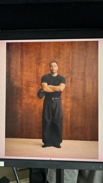 Full-body portrait of a man in black shirt and pants standing with arms crossed against a wooden backdrop.