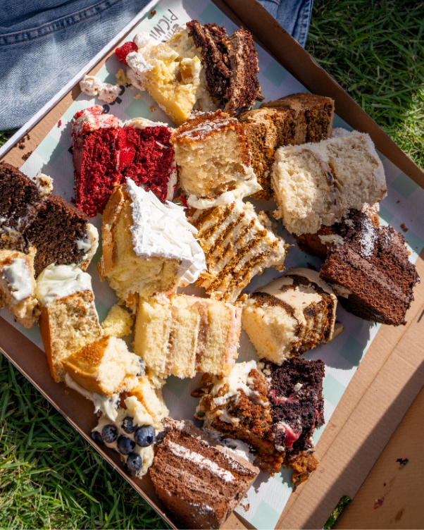 Assorted cake slices in a box on grass, featuring red velvet, chocolate, vanilla, and cream-filled varieties.