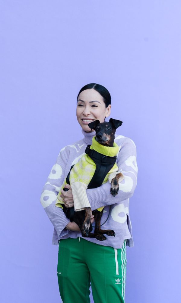 Woman in a lavender sweater holding a small black-and-tan dog in a bright yellow sweater against a purple background.