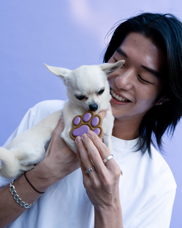 Woman smiling while holding a small white dog that smells a purple paw-shaped cookie against a lavender background, showing affection to the pet.