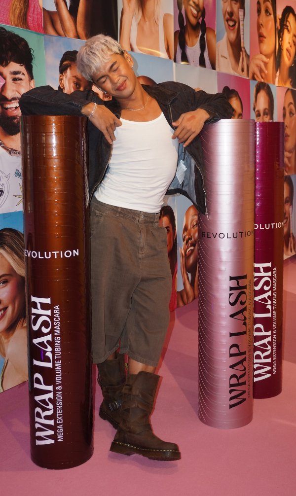 Person with platinum blond hair poses between three oversized mascara tubes in brown, pink and magenta, in a colorful cosmetics display backdrop.