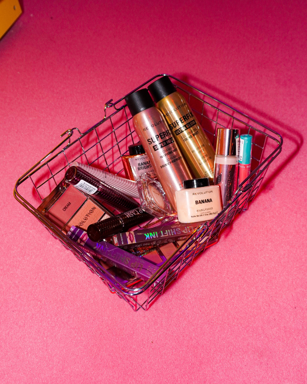 Makeup essentials in a metal basket on a bright pink surface, including powders, lipsticks, and nail polish tubes.",