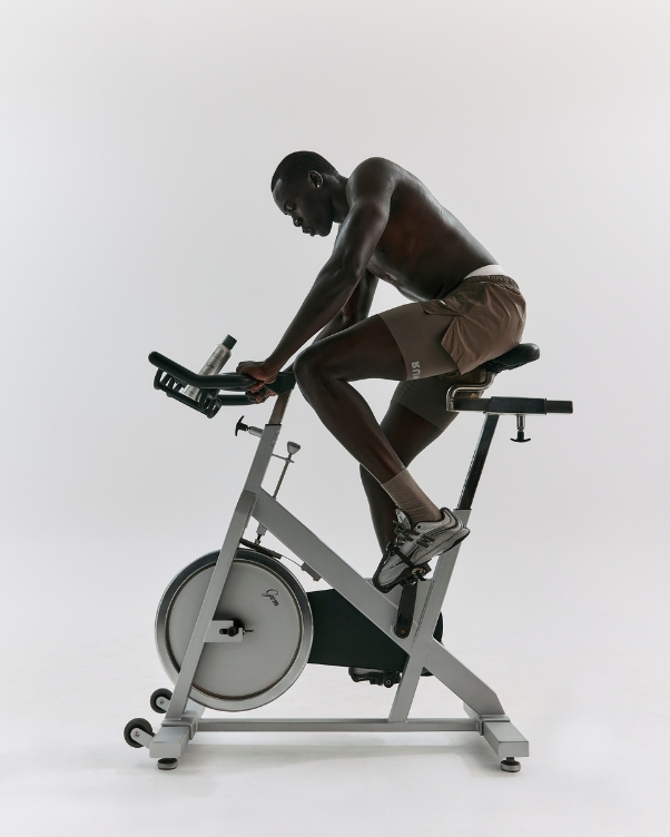 Man pedaling a stationary bike in a studio, torso bare and wearing brown shorts.