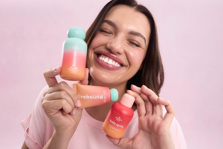 Smiling woman holding pastel orange–pink skincare bottles labeled 'rebound' against a pink background.