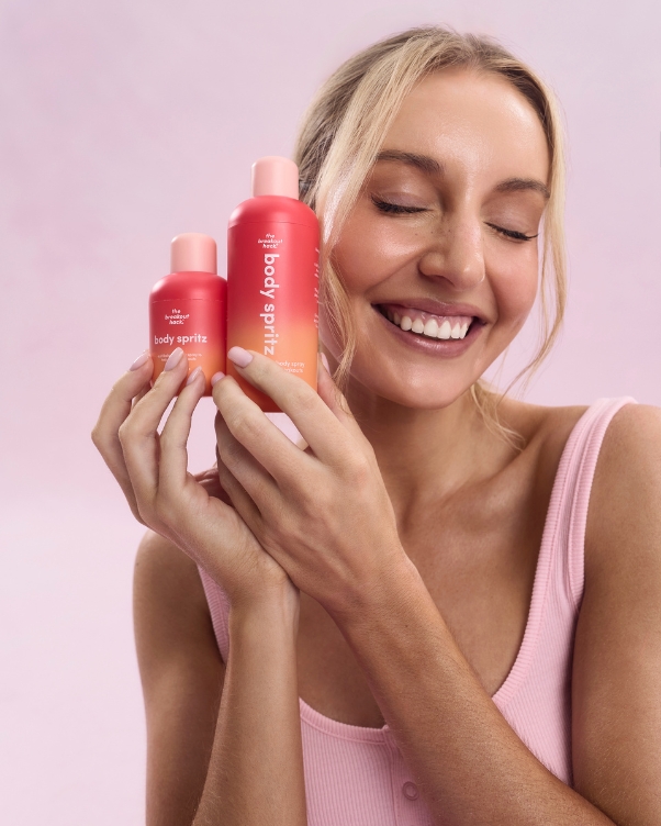 Smiling woman in a pink top holds two red body spray bottles against a soft pink background, showcasing the products.