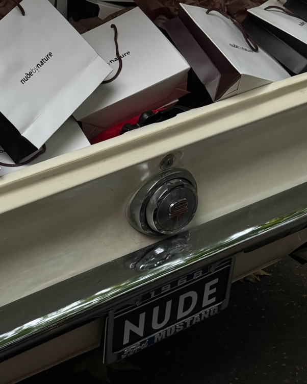 Open car trunk filled with white shopping bags, some brown bags peeking out; license plate reads 'NUDE MUSTANG'