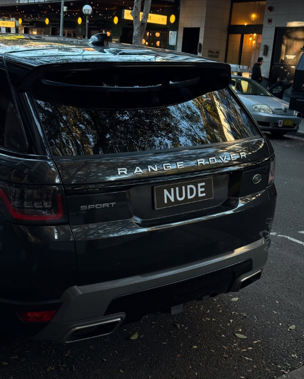 Rear view of a dark Range Rover Sport on a city street; license plate reads NUDE, reflections of trees on the rear window.