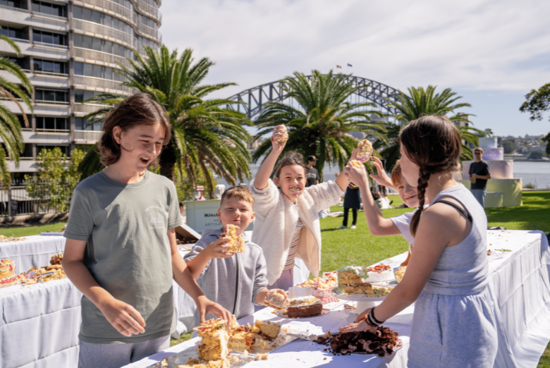 Children at an outdoor bake sale or party grab pastries from a long table, with palm trees and a water view in the background.