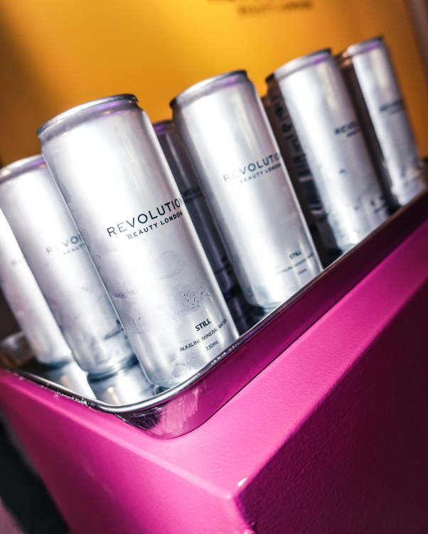Silver aluminum cans labeled 'Revolution Beauty London' in a pink display cooler; product cluster on a tilted shelf.