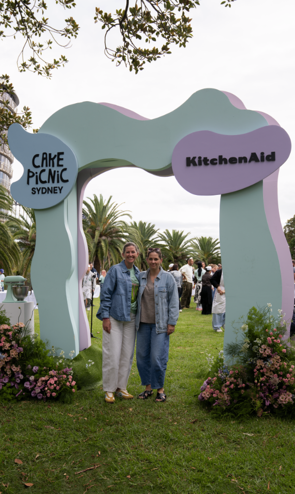 Two women pose under a pastel arch at an outdoor event labeled 'Cake PicNic Sydney' and 'KitchenAid'.