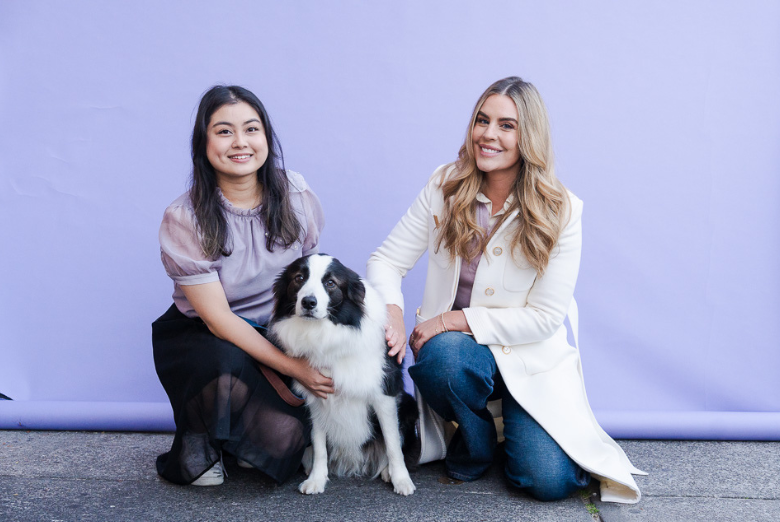 Two women kneel beside a black-and-white dog against a lavender backdrop, smiling at the camera.