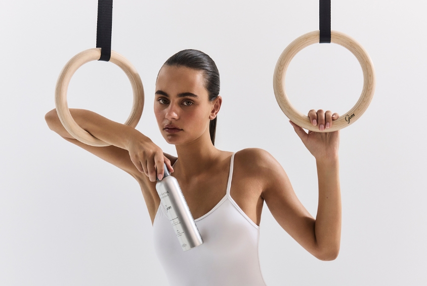 Woman in a white tank top holding a silver spray bottle between gymnastic rings in a studio setting.
