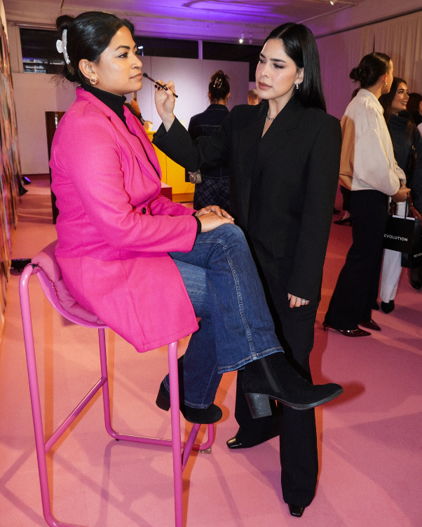 Makeup artist applies makeup to a woman in a bright pink coat seated on a pink stool at a fashion event.
