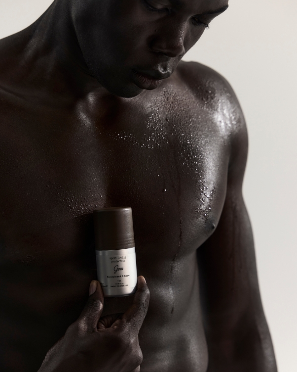 Dark-skinned, shirtless man with water droplets on his chest holds a small grooming bottle against his chest.