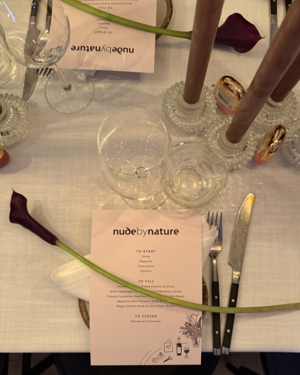 Table setting with a 'nude by nature' menu card, a purple calla lily across a white napkin, crystal candle holders, glasses, and cutlery on a white tablecloth.