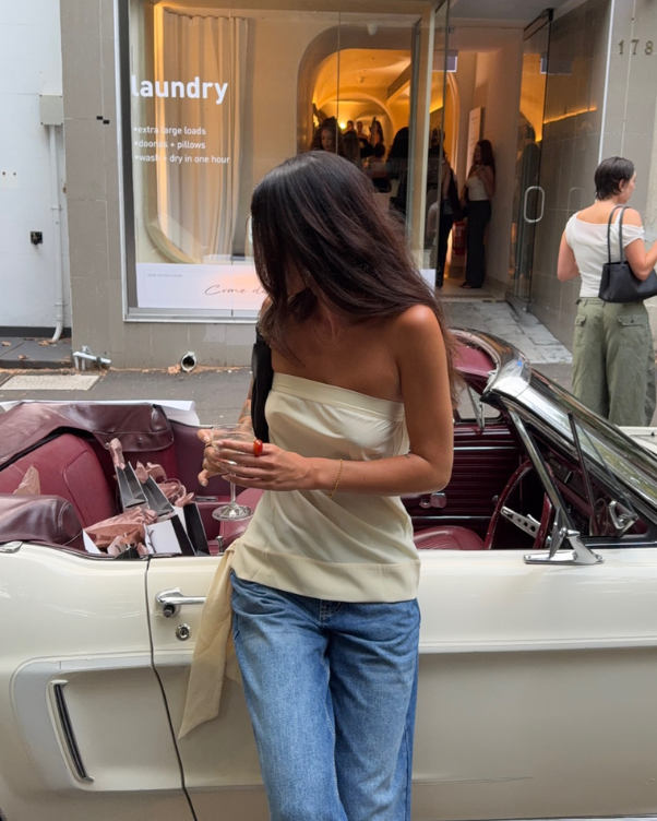Woman in a strapless cream top and jeans stands beside a white convertible, holding a glass of pink drink in front of a storefront window that says 'laundry'.