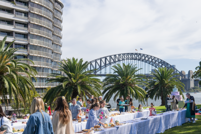 Outdoor buffet setup along a white cloth table under palm trees, with people serving and socializing, the Sydney Harbour Bridge in the distance.