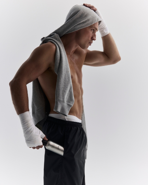 Male athlete with a gray hoodie draped over his shoulders, hand wraps, and a silver water bottle, standing in a post-workout pose.
