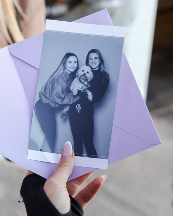 Two women pose with a small dog, holding a photo in front of a light purple envelope.