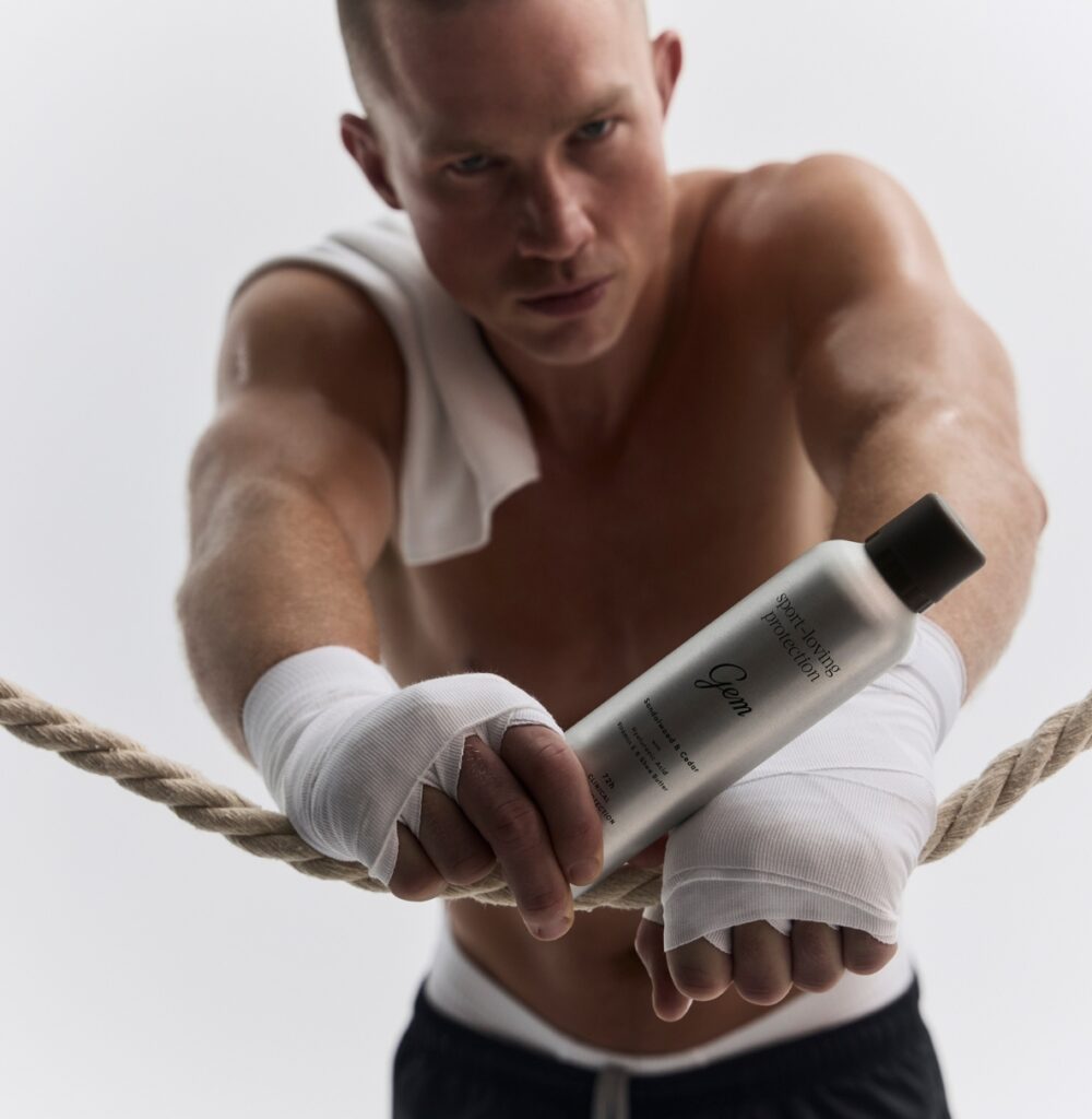 A muscular shirtless man with hand wraps grips a silver bottle labeled 'Sport-loving Protection' while leaning on a rope during a workout, against a pale background.