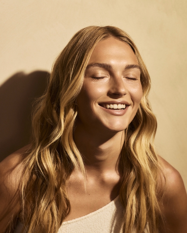 Woman with long blonde hair smiling with eyes closed against a warm beige backdrop.