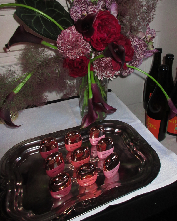 Floral arrangement in a glass vase on a table, with a silver tray of small pink-labeled bottles in the foreground.