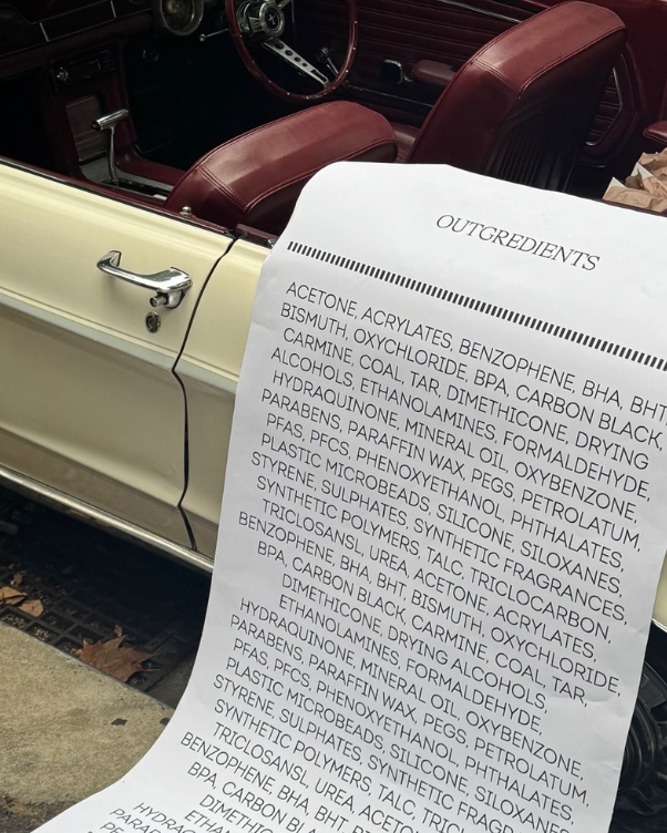 Open-door vintage cream car with burgundy seats; a long sheet labeled 'OUTGREDIENTS' listing chemical ingredients rests on the car door frame.