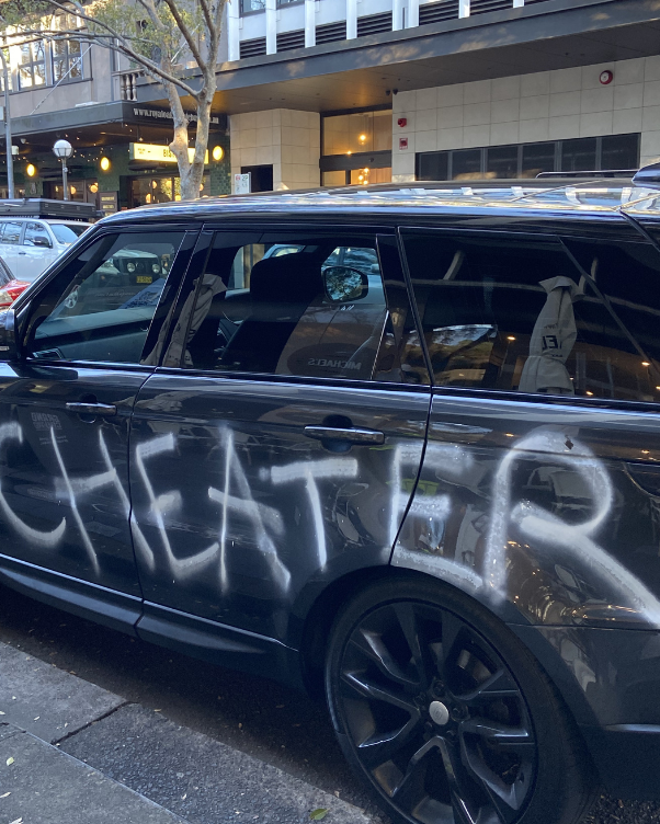 Dark SUV with large white graffiti on its side, parked on a city street with storefronts and trees in the background.