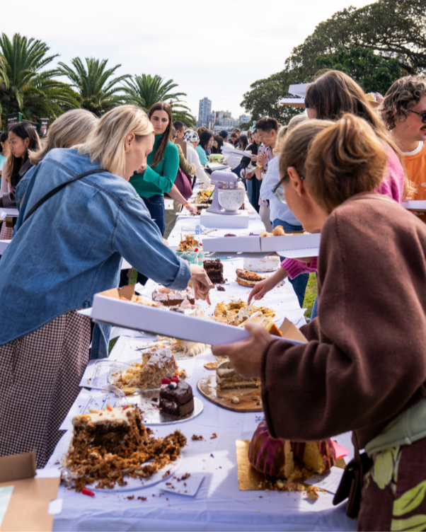 Outdoor bake sale with people selecting assorted cake slices from a long dessert table in a park setting.