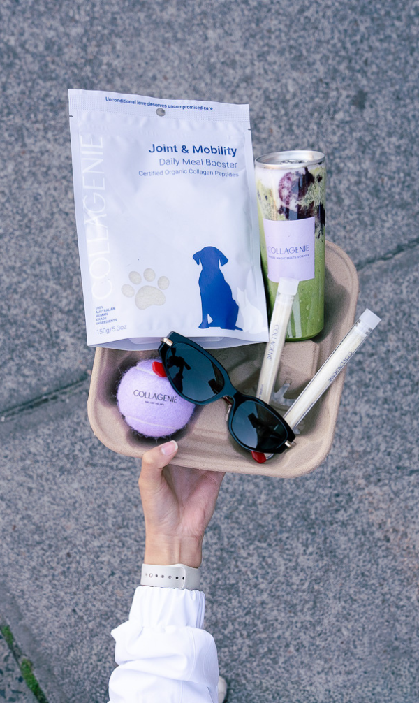 Hand holding a tray with collagen supplement pouch, bottle, sunglasses, purple ball, and two small tubes on a gray pavement background.