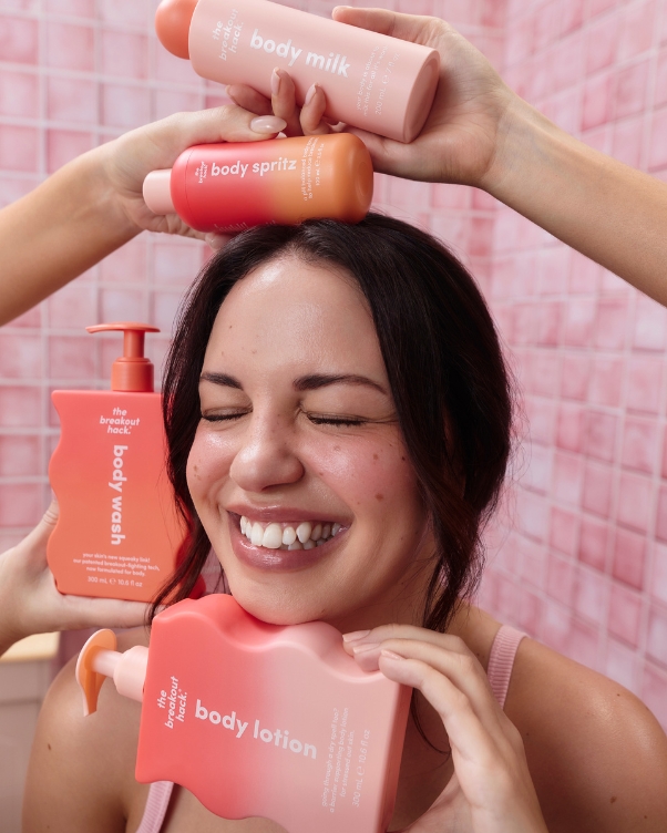 Woman smiling with pink skincare bottles around her head and shoulders, posing with rosy backdrop and products labeled body wash, lotion, and sprays.