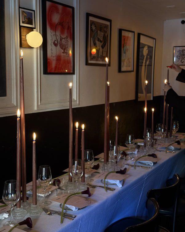 Long banquet table set for a formal dinner with tall burgundy taper candles, glassware, and neatly folded napkins in a gallery-like room with framed art on the walls.