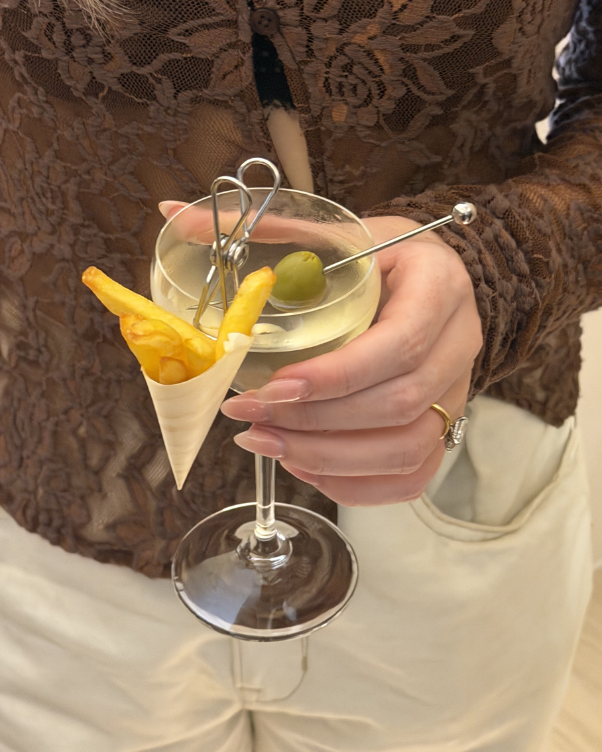 Hand holding a cocktail in a coupe glass with an olive on a pick and a cone of fries perched on the rim.