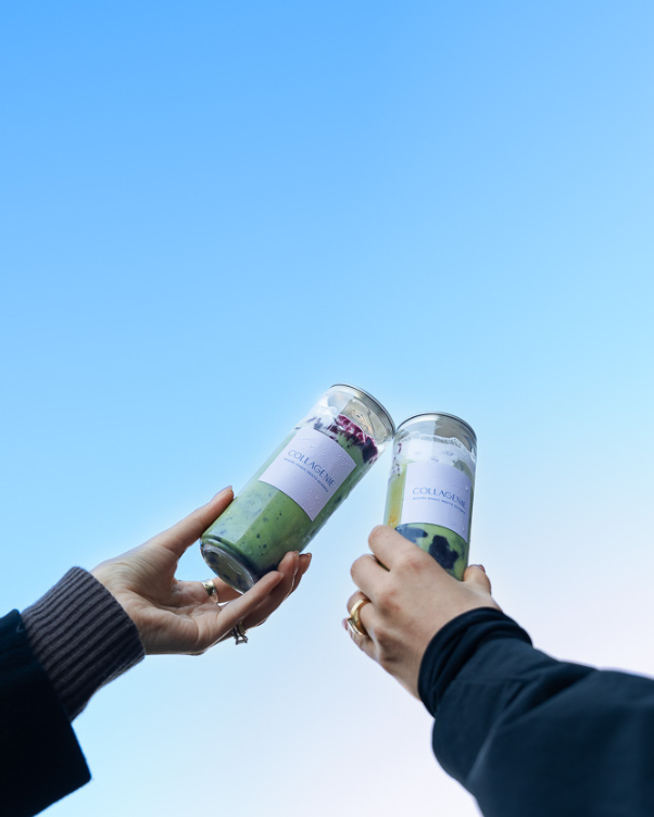 Two hands clinking green smoothie bottles labeled Collagen against a bright blue sky in the background.