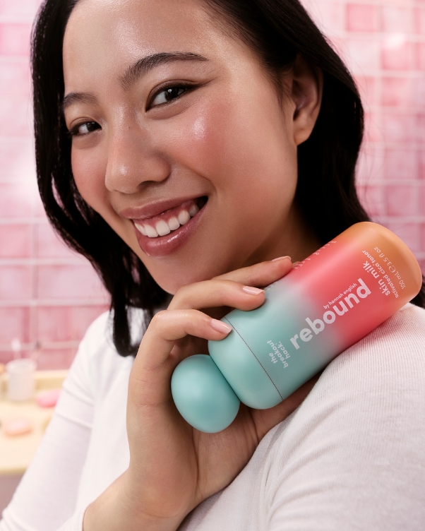 Smiling woman holds a pastel gradient bottle labeled 'rebound' against a pink tiled background.