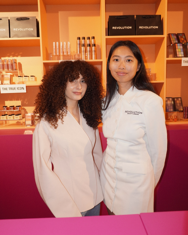 Two women in white coats pose together in a cosmetics shop with orange shelves and Revolution products behind them.