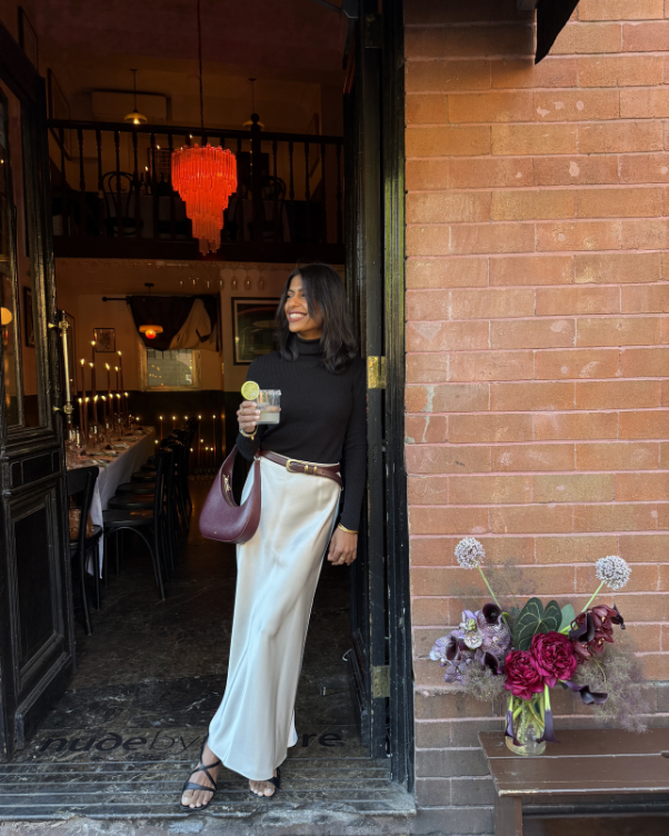 Woman standing in restaurant doorway, wearing a black top and ivory satin skirt, holding a lime-garnished drink with a purple handbag at her side.