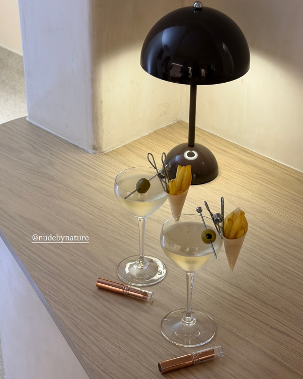 Two pale cocktails in coupe glasses with olive skewers and cones of fries on a light wooden table, under a black desk lamp placed at the back.