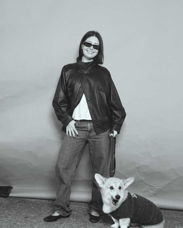 Woman in a black leather jacket, jeans, white tee and sunglasses posing with a corgi wearing a knit sweater in a studio setting.