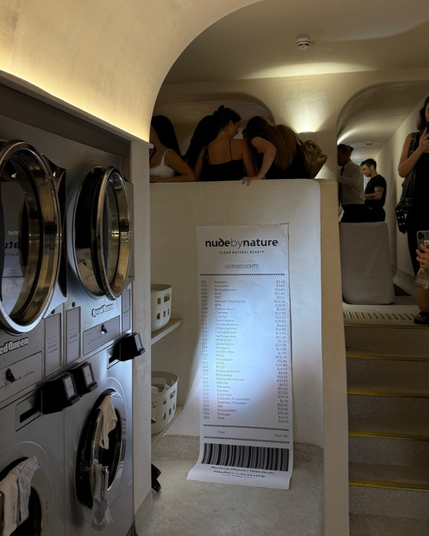 Laundromat-like space with front-loading washers on the left, a tall receipt poster in the center, and people standing on a curved balcony in the background.