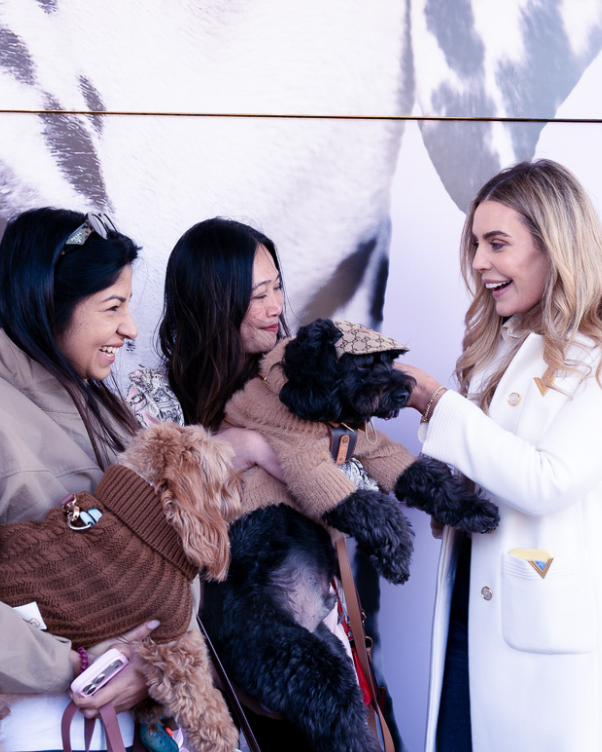 Three women smile and hold fluffy poodles dressed in sweaters at a pet event.
