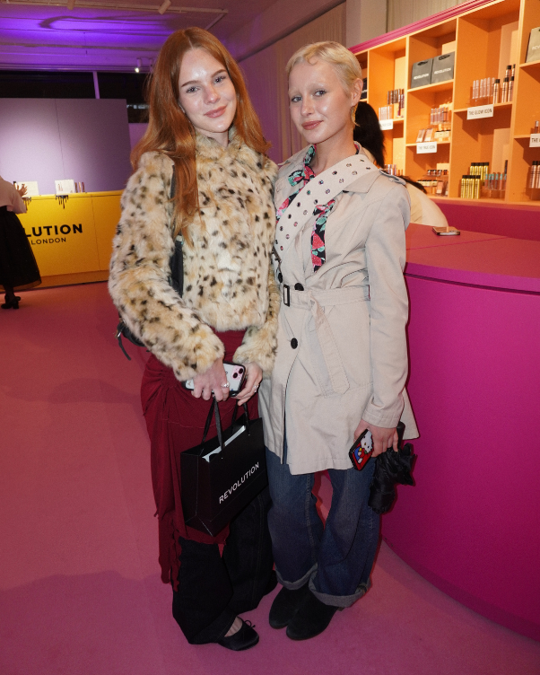 Two women pose together in a bright pink retail space, one in a faux fur coat and red hair, the other in a beige trench coat with a scarf and jeans.