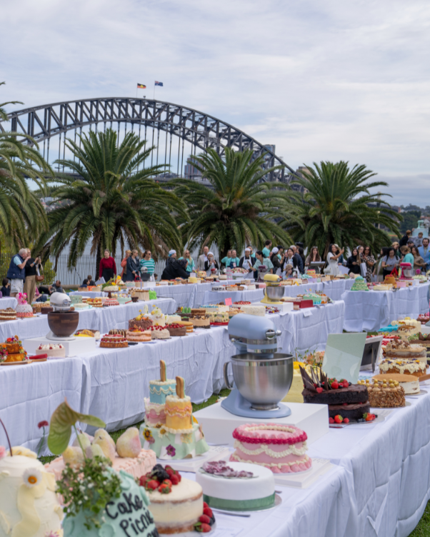 Outdoor cake display along a waterfront with white-clothed tables, many decorated cakes, and a crowd browsing; palm trees and a large bridge in the background.
