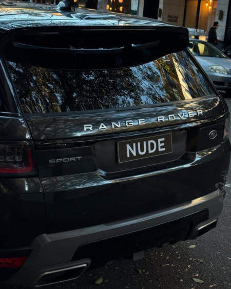 Rear view of a black Range Rover Sport parked on a city street, license plate reads 'NUDE' with reflections of trees in the rear window.