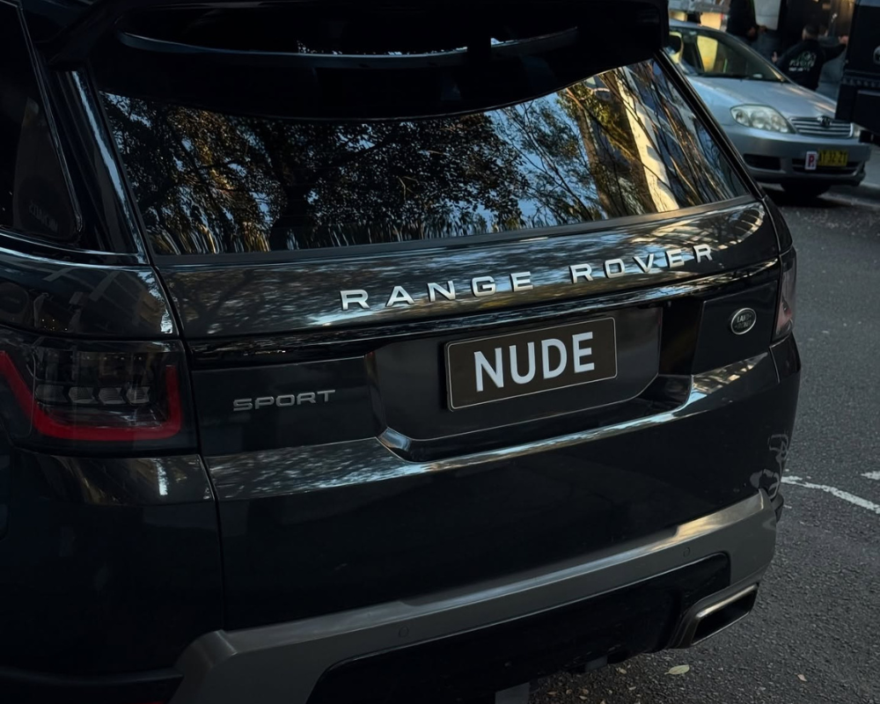 Rear view of a black Range Rover Sport parked on a city street, license plate reads 'NUDE' with reflections of trees in the rear window.