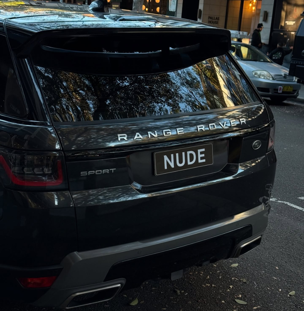 Rear view of a black Range Rover Sport parked on a city street, license plate reads 'NUDE' with reflections of trees in the rear window.