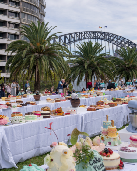 Outdoor cake festival with dozens of decorated cakes on white-clothed tables, palm trees, and a steel arch bridge in the background.